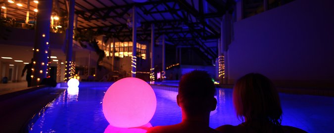 Ein Paar sitzt am Rand eines großen, blau beleuchteten Schwimmbeckens, auf der Wasseroberfläche treiben bunte Leuchten. Es ist weitgehend dunkel, nur im Hintergrund sind wenige Lichter und Kerzen zu sehen.